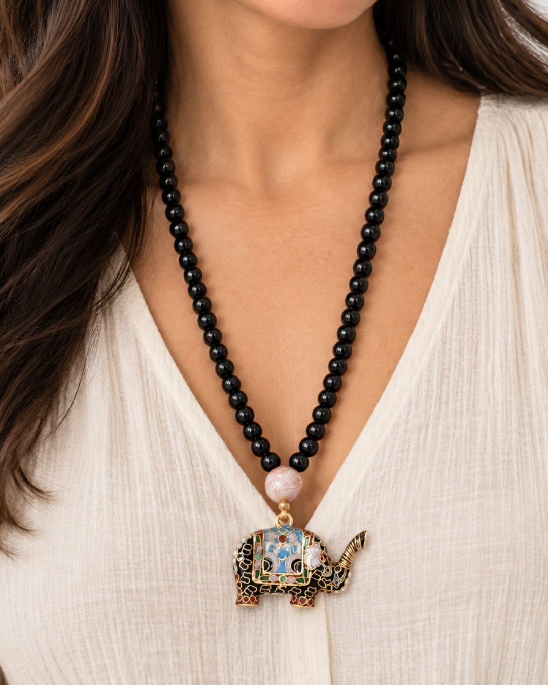 Black stone bead necklace with cloisonne elephant pendant worn by a model in a white linen top, showing length and scale